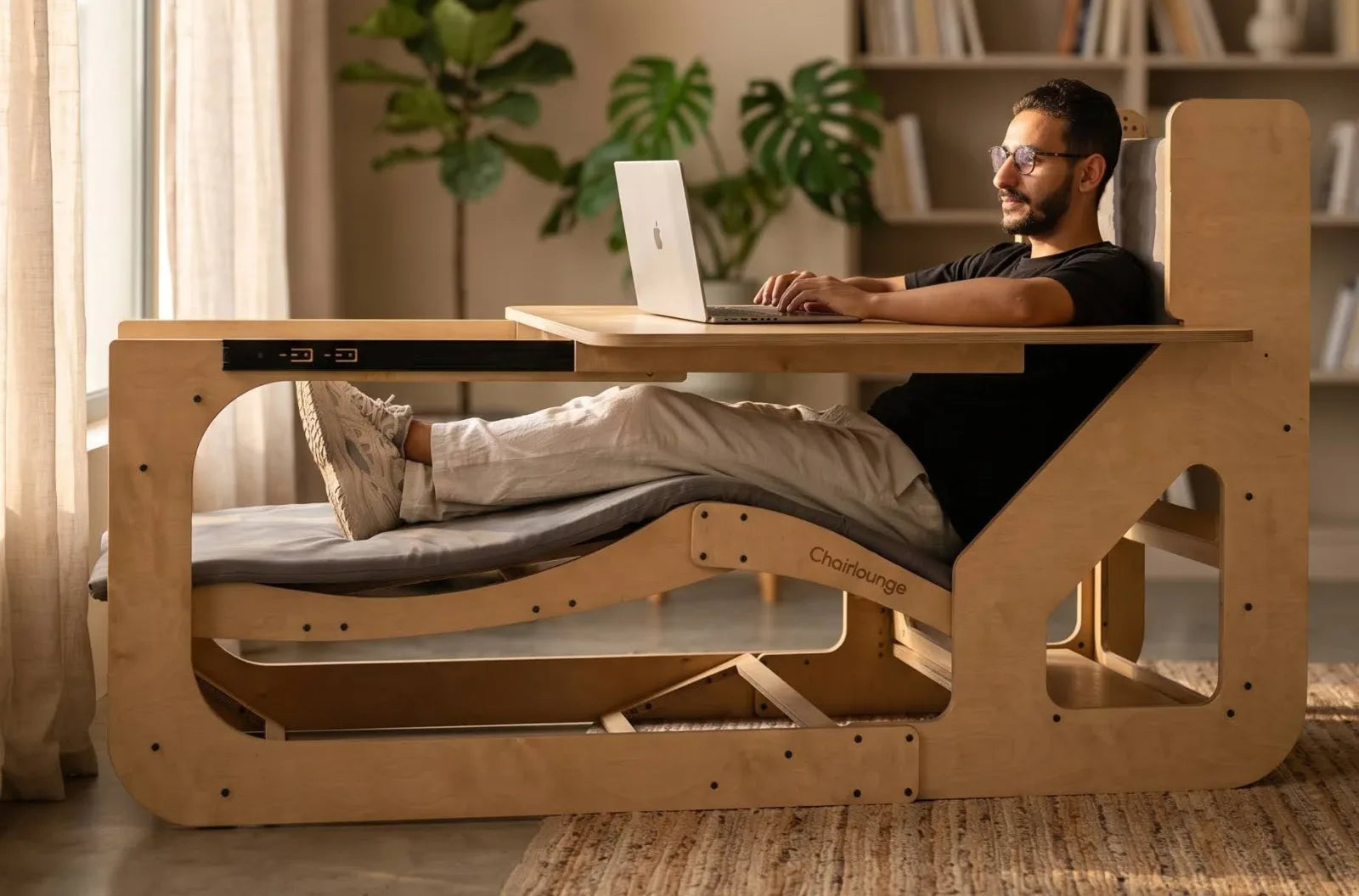 رجل مستلقٍ على ChairLounge وساقاه مرفوعتان يعمل على لابتوب فوق المكتب المنزلق